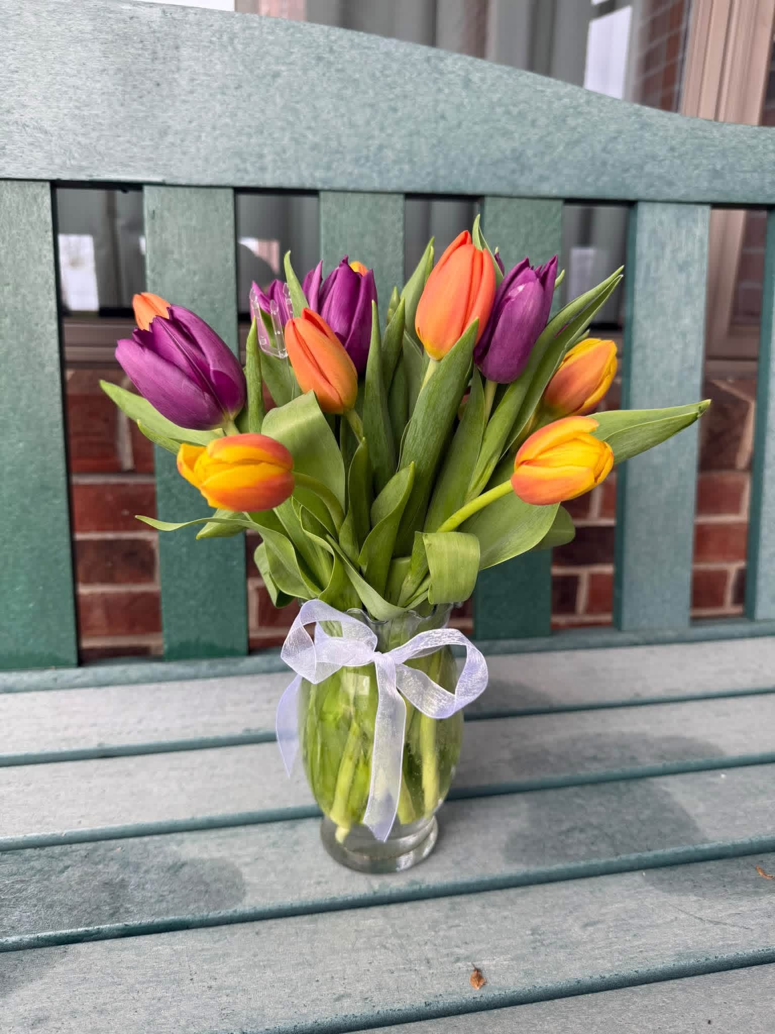 Purple and orange tulips with white ribbon bow in glass vase on garden bench