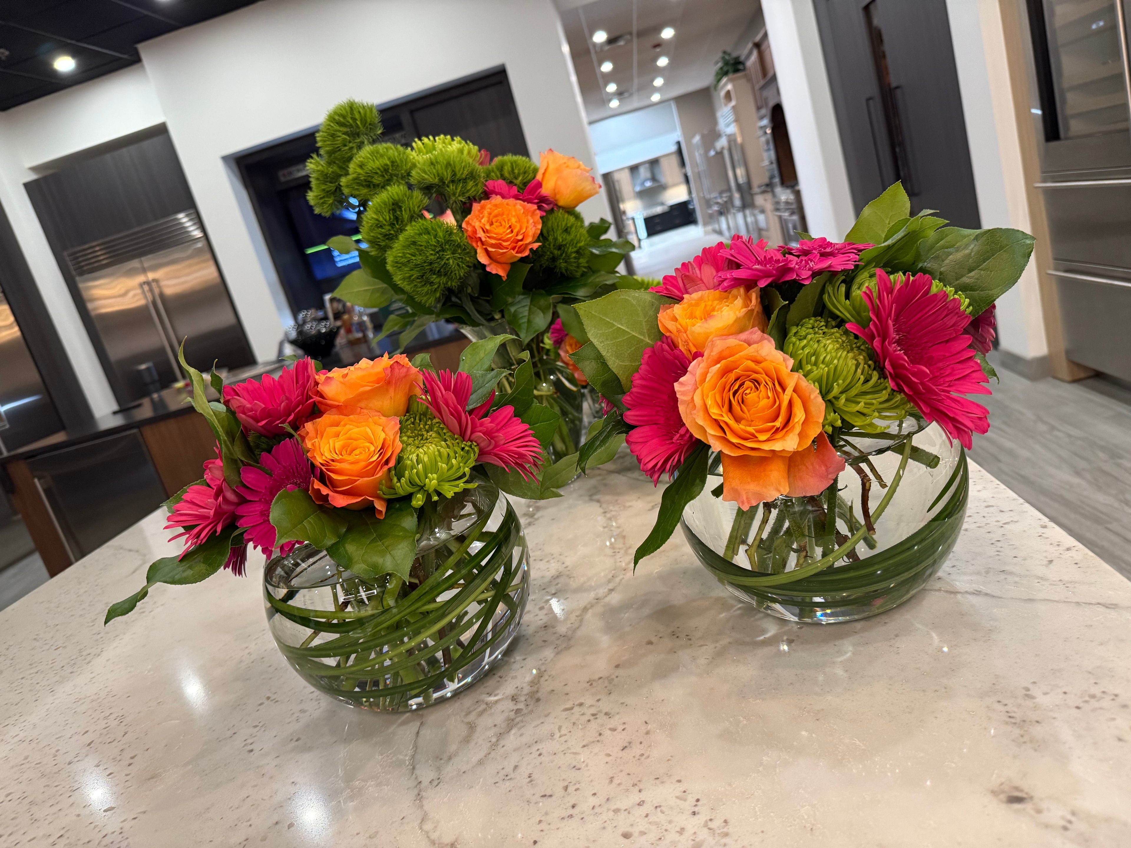 Two round glass bowl arrangements with orange roses, pink gerberas, and green chrysanthemums